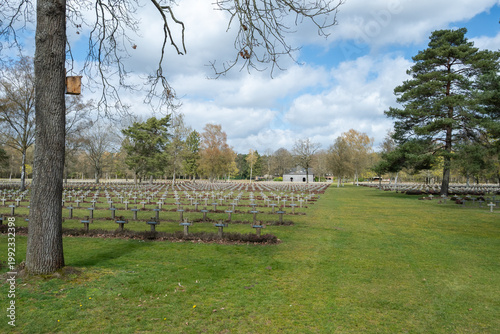 German War Cemetery Lommel. A cemetery landscape with rows of tombstones and a cloudy sky, situated in a serene location with abundant greenery.
