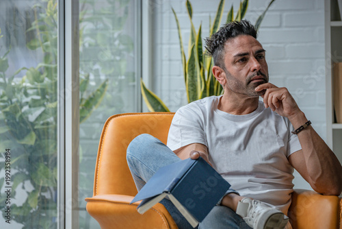 Fotografi Middle-aged man in casual attire pausing from reading, gazing pensively into the