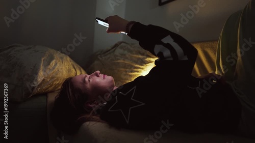 Girl doom scrolling her smartphone on a coach at night. Social network and phone addiction concept. Cozy home ambient light