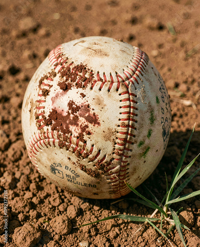 A close-up of an old baseball resting on the ground with some grass in the foreground. Baseball stitching macro dirty ball.