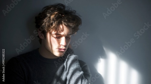 Sad young man sitting alone in moody light, dramatic shadows on face, copy space on grey wall background for mental health or loneliness concepts