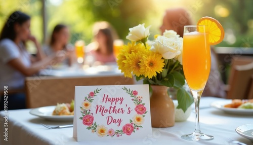 Table set for Mother Day brunch outdoors. Features a mimosa cocktail, flower bouquet, and greeting card. People dine in background, enjoying sunny day.