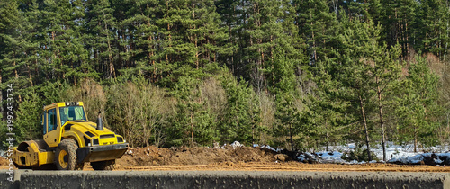 Yellow road roller creating a new roadbed against a backdrop of a pine forest, showing the ongoing process of land development and construction work in a natural setting.