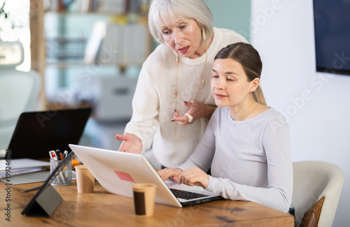 Elderly man helps young female colleague use laptop while working in office space