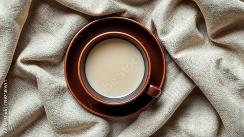close cup coffee beige fabric background cup placed center saucer slightly overlapping saucer dark brown color has handle one side coffee cup filled light colored liquid which appears creamy white