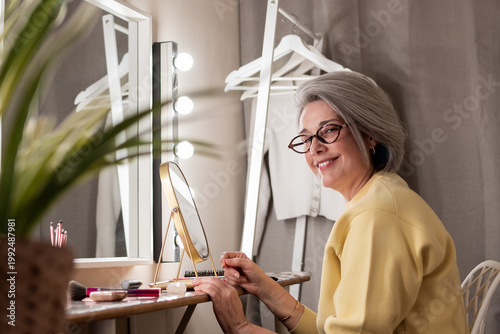 Senior woman in glasses smiles at camera seated at a vanity with mirror and makeup, enjoying a joyful self-care and beauty routine at home, calm, confident and content