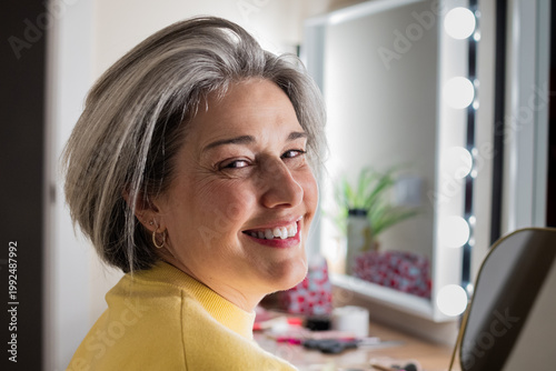Smiling senior woman with gray hair looking back at the camera from her vanity table, embracing natural aging and enjoying a moment of self-care and positivity in her home