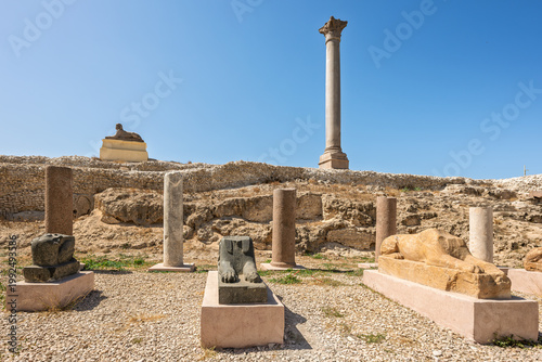 The ancient Roman monolithic column rises above Egyptian sphinx figures and ruins at the archaeological site of Pompey's Pillar in Alexandria Egypt.