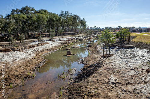 A newly constructed urban wetland and creek restoration project under development in a suburban Melbourne, Australia. Freshly planted native saplings, placed boulders,  muddy soil, and a shallow water