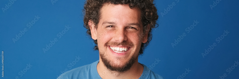 Fototapeta premium Close up, guy with curly hair dressed in blue t-shirt looks at the camera with smile isolated on blue background in the studio. Panoramic. Panoramic