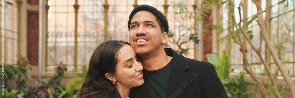 Fototapeta premium Happy young couple embracing in greenhouse, with background of arched windows and plants. Panoramic