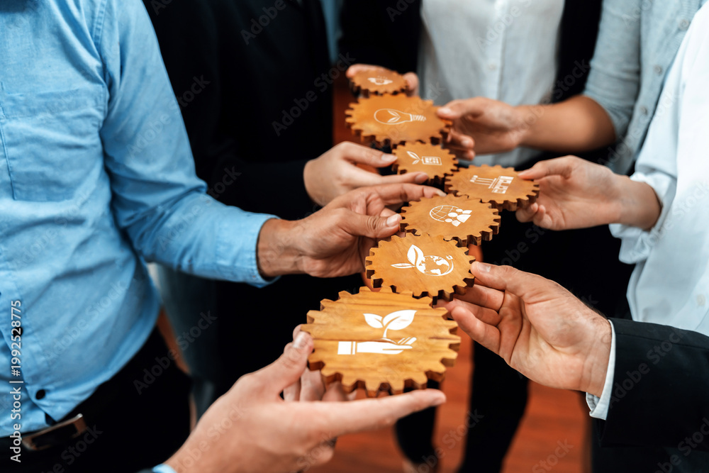 Naklejka premium A close-up view of diverse hands holding wooden gears, each marked with eco-friendly symbols. This image showcases teamwork in promoting sustainable business practices and growth. Amity