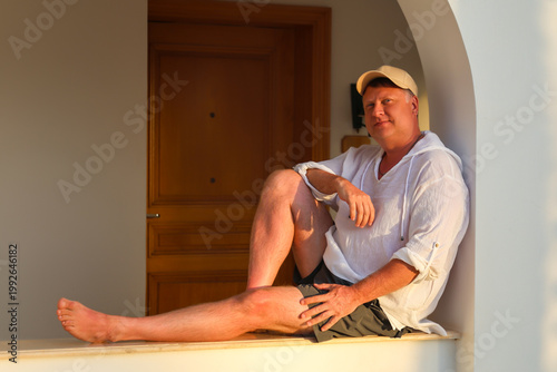 Adult man in a white shirt and cap sitting on a wide windowsill, enjoying the beautiful sunset, reflecting on life and tranquility in the golden hour.