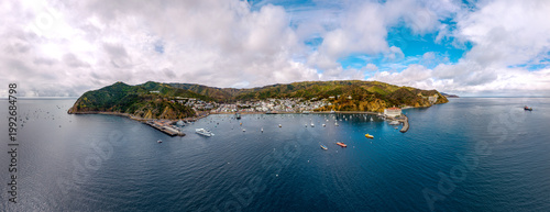 Aerial view of a narrow peninsula town surrounded by deep blue water, green hills, scattered buildings, and boats moving through the harbor.