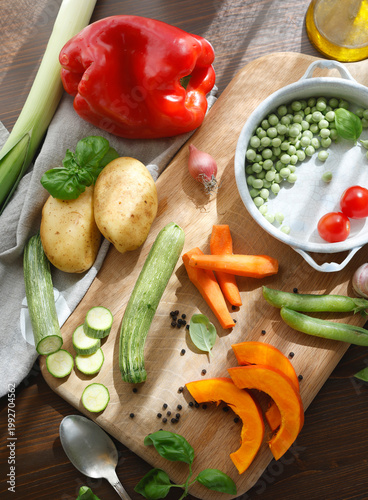 Assorted organic vegetables and vintage utensils on wooden board in sunlit countryside kitchen. High-angle shot captures sliced pumpkin carrots potatoes and zucchini with fresh green peas in ceramic