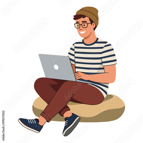 A smiling man sits cross-legged on a beanbag chair working on his laptop