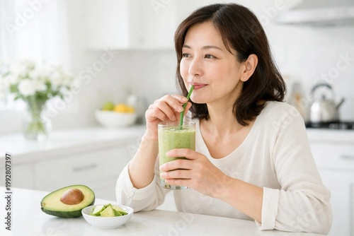 Healthy middle-aged Asian woman drinking a green avocado smoothie in a bright modern kitchen.