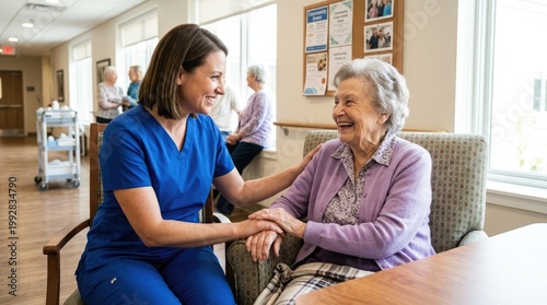 A smiling nurse sits and talks with a laughing elderly woman in a comfortable lounge area. The nurse is wearing a blue uniform and the woman is seated in a patterned armchair.