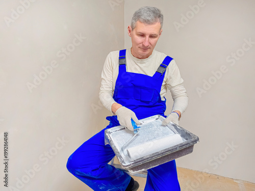Gray-haired man paints wall with roller, wearing blue overalls.