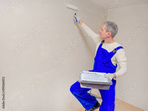 Man paints wall with roller, wearing blue overalls