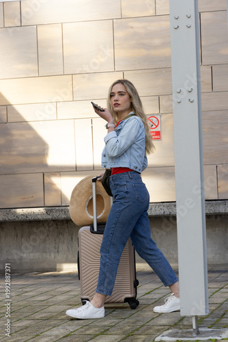 Woman walking across plaza pulling rolling suitcase with straw hat and speaking into phone