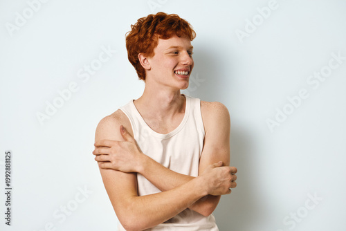 Foto Young man with red hair in white tank top smiling and hugging himself on light background