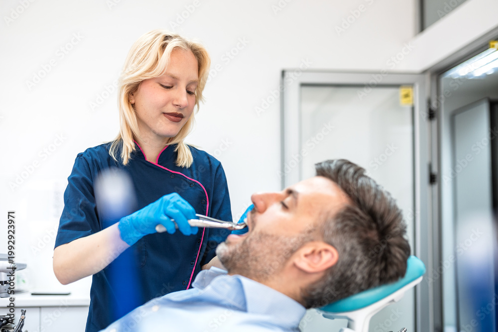 Fototapeta premium Female physician performing tooth extraction for male patient with dental pliers. Blonde woman doctor conducting oral surgery in modern clinic. Healthcare and medicine professional dental service.