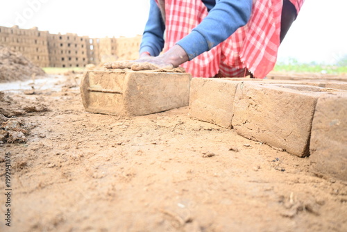 A person manually shaping bricks in an open field. Using a rectangular wooden mold and raw, reddish-brown clay, the worker skillfully forms each brick on the dusty ground. Slow motion video.