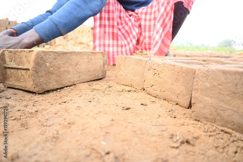 A person manually shaping bricks in an open field. Using a rectangular wooden mold and raw, reddish-brown clay, the worker skillfully forms each brick on the dusty ground. Slow motion video.