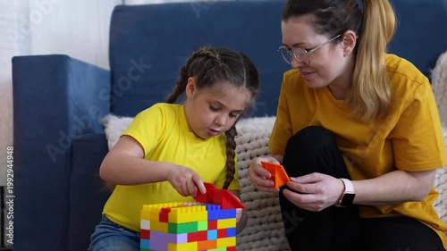 Mother and daughter build block tower on couch while child arranges colorful toy brick and parent guides play and learning in family living room with focused attention and joyful interaction