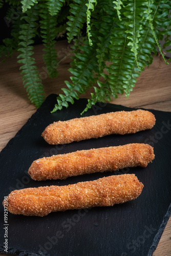 Sticks of cheese are coated in breadcrumbs and fried until crispy, arranged on a black plate. Overhead shot.