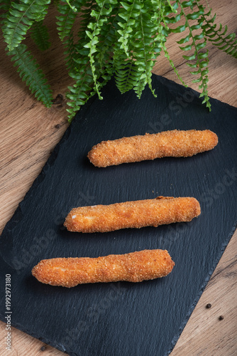 Sticks of cheese are coated in breadcrumbs and fried until crispy, arranged on a black plate. Overhead shot.