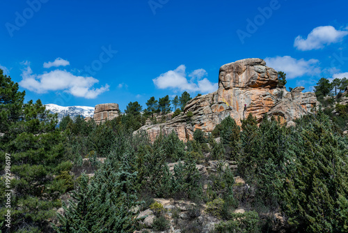 Wide landscape view of La Pedriza featuring a granite rock formation with a barely visible climber in the distance, emphasizing scale and vastness. Surrounded by forest and with a snow-capped mountain