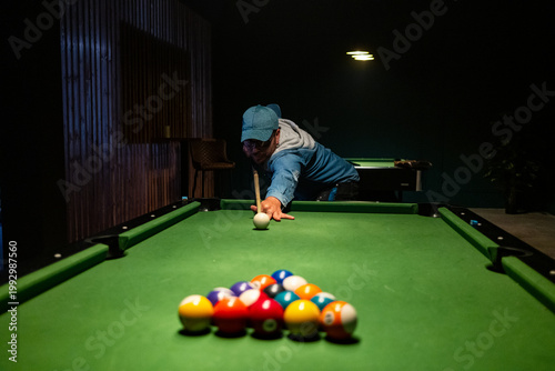 Man in a cap taking a shot on a green pool table in a dimly lit bar