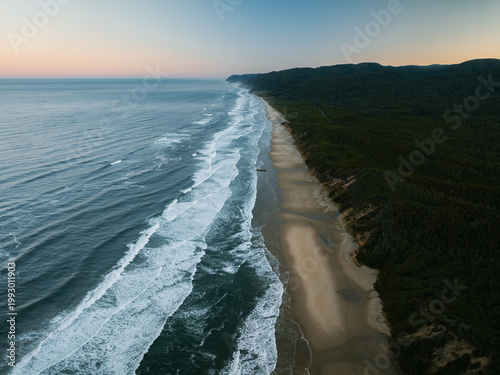 Aerial view of the Oregon Coast Highway winding through dense evergreen forests along a sandy beach with crashing waves at dusk in Florence, Oregon, United States.