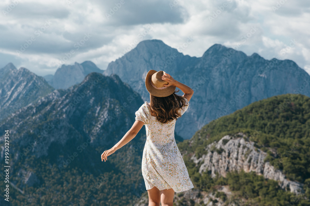 Fototapeta premium Rear view of a young female tourist in a summer dress standing on a cliff edge. She is holding her hat against the wind while admiring the breathtaking panoramic view of the rocky mountain landscape.