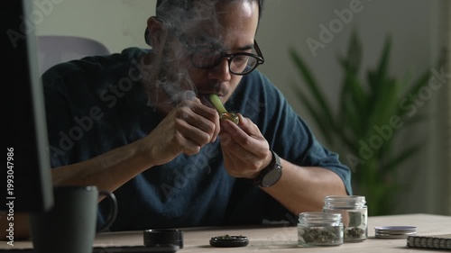 Man smoking a cannabis from smoking pipe, with cannabis equipment and accessories on the table.