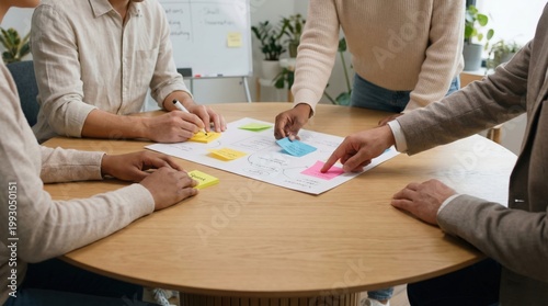 Group of diverse individuals collaborating around a round wooden table, using colorful sticky notes and discussing ideas on a large sheet of paper in a well-lit office environment