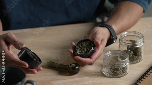 Man using cannabis grinder, with cannabis equipment and accessories on the table.