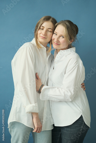 Beautiful mature mother and adult daughter in white shirts and jeans hugging in the photostudio on blue paper background. Two female generations. Mother's day concept
