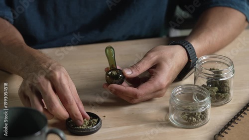 Man filling glass pipe with cannabis, with cannabis equipment and accessories on the table.
