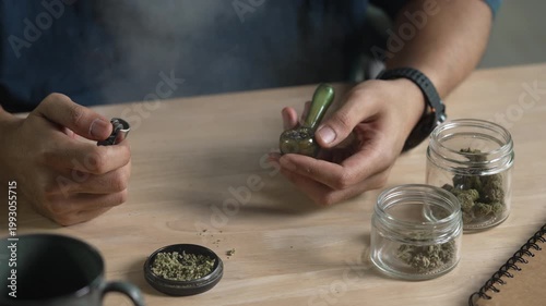 Man smoking a cannabis from smoking pipe, with cannabis equipment and accessories on the table.