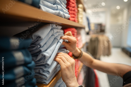 Female purchasing new basic clothing at garment fashion showroom, woman looking through stack of t-shirts on shelf display, examinees quality of material, fabric and preferable size of clothing item.