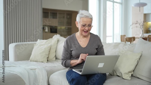 Confident stylish happy middle aged senior woman using laptop at home. Stylish older mature 60s grandmother sitting at couch looking at computer screen typing chatting reading writing email