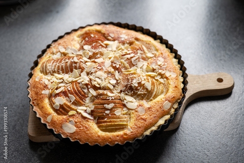 Baked pear tart with almond topping sits on a wooden board in a kitchen setting during the day