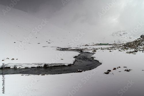 A minimalist winter landscape of Zero Point in North Sikkim, featuring a dark, winding river cutting through a vast, pristine expanse of pure white snow under high-altitude Himalayan conditions.