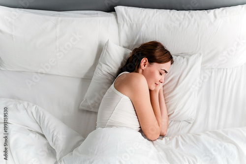 Fotografía A woman sleeps peacefully on her side in a white, minimalist bed with fluffy pillows and a cozy blanket, creating a calm, restful bedroom scene