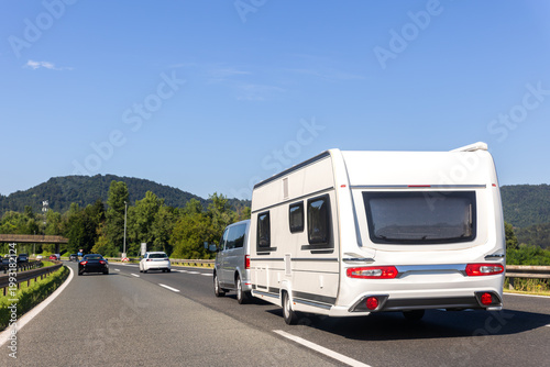 Scenic view big modern white family car tow rv camper vehicle drive Europe highway road freeway drive cloud in summer day. Rving motorhome lifestyle travel and adventure tourism trip journey concept