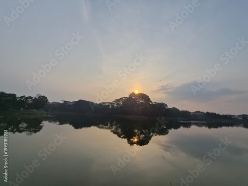 Obraz na plátne Serene sunrise over a tranquil lake with trees reflecting in the calm water under a soft, cloudy sky