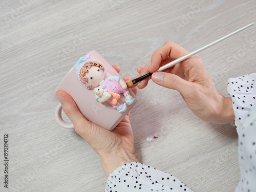 Hands paint a pink mug with a tiny doll design